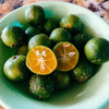 A green bowl filled with many small, green calamansi fruits, with one fruit cut in half to show the orange-colored interior.