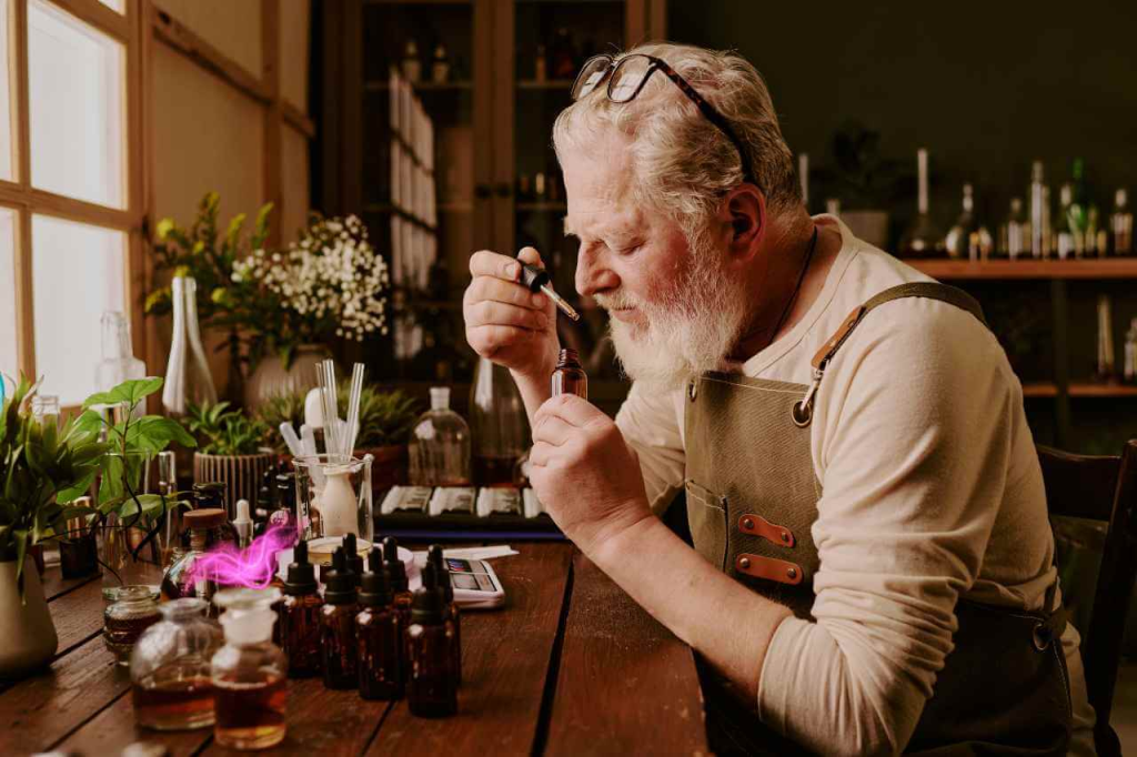 A senior man with a white beard and glasses wearing an apron, smelling a small brown glass bottle with a pipette in his hand, in a workshop filled with various bottles and greenery.