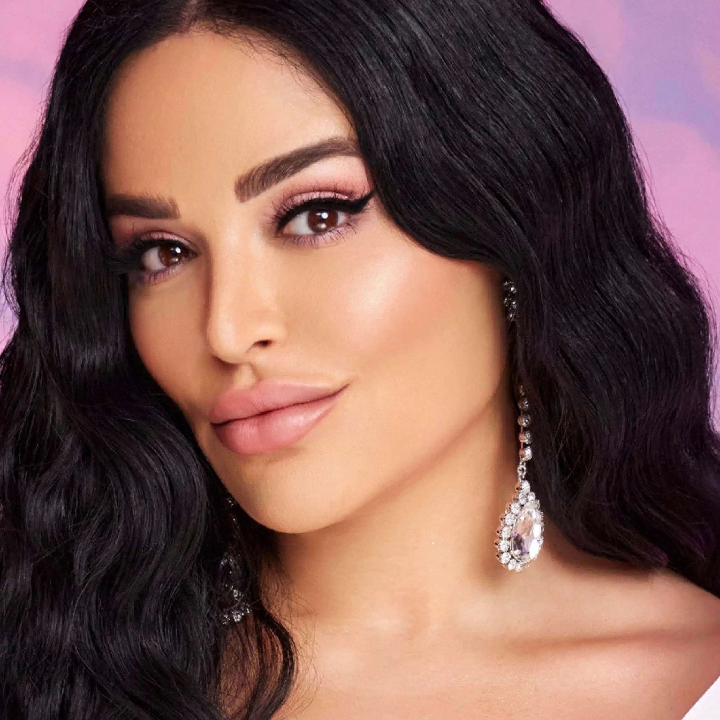 Close-up portrait of Mona Kattan with long black wavy hair, glamorous makeup, and a statement crystal earring, set against a soft pink background.