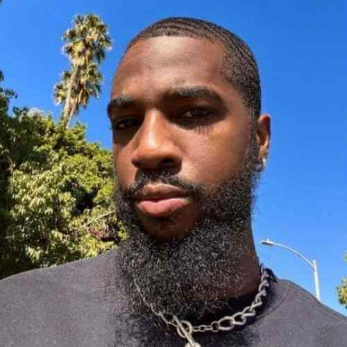A close-up outdoor portrait of a man with a beard and short hair, wearing a silver chain necklace.