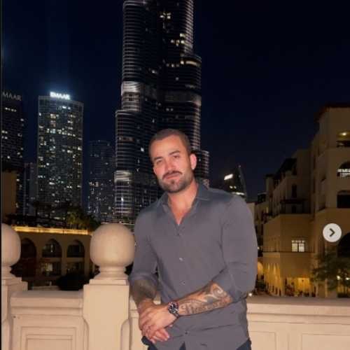A man with a beard and tattoos stands on a balcony at night with the illuminated Burj Khalifa in the background.